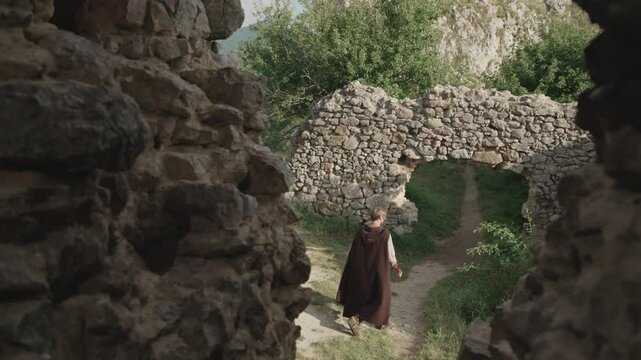 Viewed through a gap in a stone wall, a man in a historical brown cloak walks along a path and through an ancient archway in the ruins of a medieval fortress.