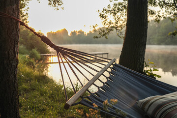 Striped pillow on a blue hammock at sunrise by a calm lake, with soft mist on the water and golden...