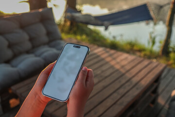 Hands holding smartphone with blank screen on wooden terrace at lakeside during sunrise. Hammock and cozy outdoor furniture in background create relaxing summer atmosphere for mockup use
