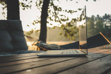 Laptop placed on a wooden table in a tranquil forest setting with a hammock and soft morning light,...