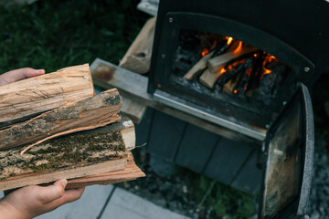Person holding chopped firewood near an outdoor black metal stove with a burning fire inside,...