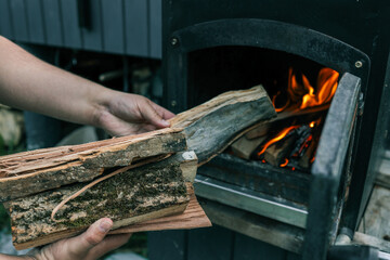 Person holding chopped firewood near an outdoor black metal stove with a burning fire inside,...