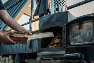 Person holding chopped firewood near an outdoor black metal stove with a burning fire inside,...
