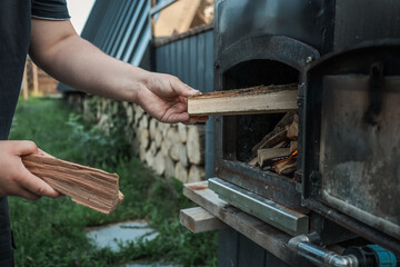 Person holding chopped firewood near an outdoor black metal stove with a burning fire inside,...