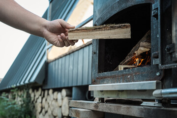 Person holding chopped firewood near an outdoor black metal stove with a burning fire inside,...