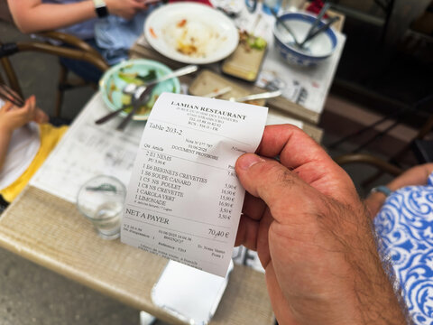 Strasbourg, France - Jun 1, 2025: Close-up of restaurant receipt held over table with Asian food plates and glass of water, outdoor terrace seating, total 70.40 euros.