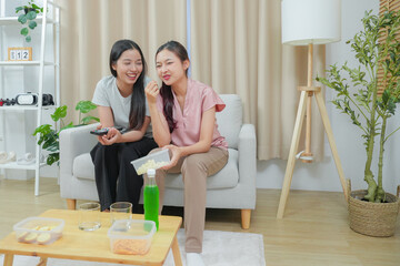 Happy friends asian woman enjoying watching television with popcorn using remote control choosing TV show movie. Enjoying television relaxing at home together.