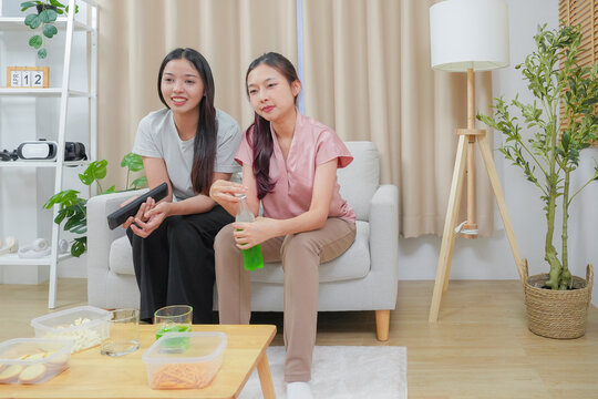 Happy friends asian woman enjoying watching television with popcorn using remote control choosing TV show movie. Enjoying television relaxing at home together.