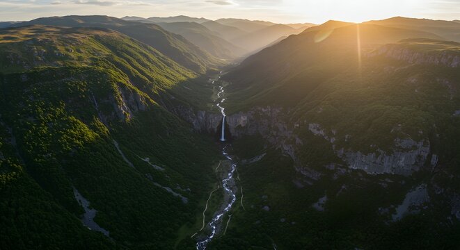 Waterfall cascades down into a deep river valley as the golden sunset light illuminates the rugged mountain peaks above.