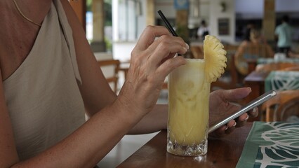 Woman holding phone and sipping tropical juice in cozy cafe enjoying peaceful moment while staying...