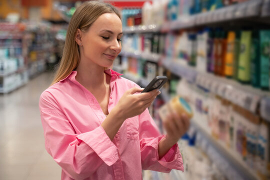 Young woman scanning product barcode with her smartphone while shopping for cosmetics in supermarket, checking ingredients and comparing prices - Powered by Adobe