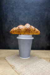 A golden brown croissant creatively placed on top of a kraft paper cup on a wooden surface with a...