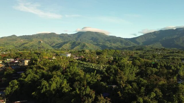 Early morning aerial ascent reveals vibrant greenery and scattered village houses below Mount Apo, with a soft cloud crowning the peak and golden sunrise illuminating Mindanao&rsquo;s landscape.
