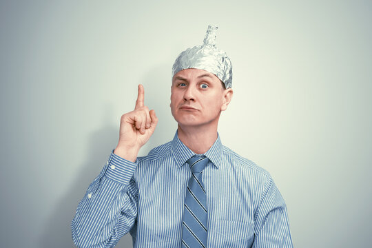 Suspicious man in striped shirt and tie wearing a DIY tin foil hat, raising finger with intense expression. Conspiracy theorist concept on light grey background.