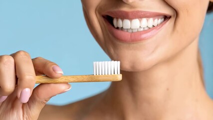 Attractive woman with healthy, clean, white teeth is holding a bamboo toothbrush against a light blue background, promoting dental hygiene and oral care products.