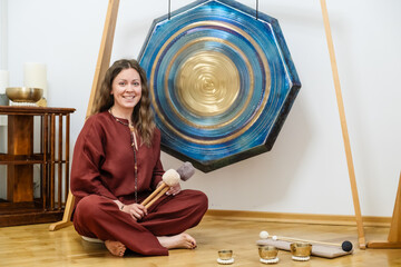 Woman sitting on the floor holding mallets, smiling warmly beside a large harmony gong. Room is set...
