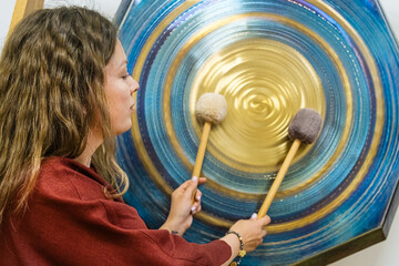 Woman plays large gong using mallets, producing harmonious sound waves. She appears focused and...