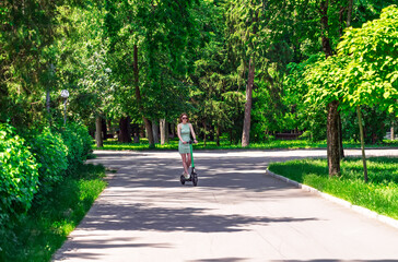 Modern urban transport: A stylish girl in a dress enjoying a ride on an e-scooter in nature.Summer leisure and active lifestyle. A woman on a kick scooter in a public city park.Eco-friendly commute