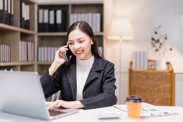 Young businesswoman is smiling while working on her laptop and speaking on the phone, sitting at...