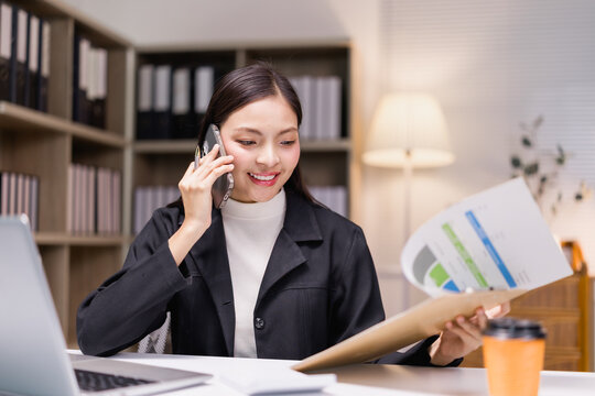 Young professional woman is working with financial documents while having phone conversation in modern office, demonstrating efficiency and multitasking skills - Powered by Adobe