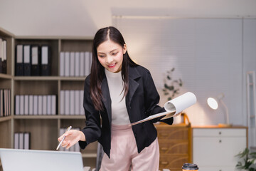 Young Asian businesswoman standing in her office, holding documents in one hand and pointing at her laptop with a pen in the other hand, smiling and working efficiently in a modern workspace