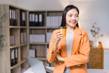 Young asian businesswoman wearing orange jacket drinking coffee from disposable cup and smiling...