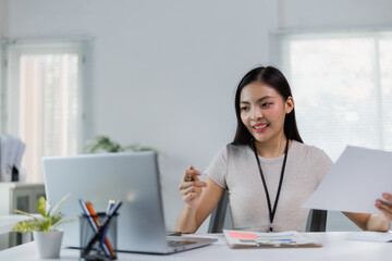 Asian businesswoman participating in a video conference on her laptop, reviewing documents while...