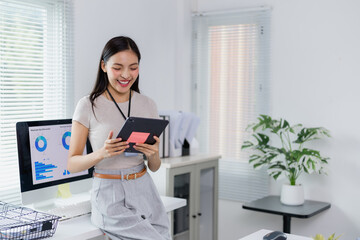Young office worker holding digital tablet and checking project statistics on computer, enjoying work in modern company office with big windows and plants