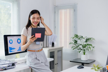 Businesswoman is using a tablet and analyzing financial data on a computer in the office, touching her hair with a thoughtful expression as she reviews the information on the screen