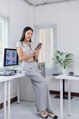 Young businesswoman wearing an id card and leaning on her desk while using her smartphone in a modern office, with a computer showing charts