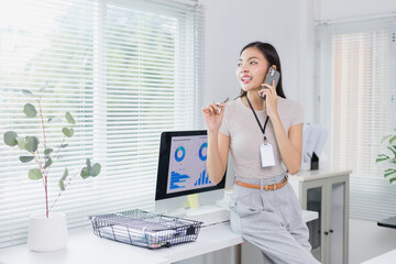 Young businesswoman wearing an id card is sitting on her desk and talking on the phone while holding a pen and looking away, with computer showing graphs in front of her in a modern office