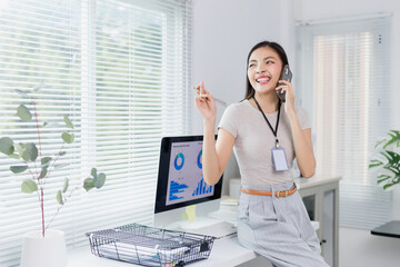 Young businesswoman wearing an id card is sitting on her desk, holding a pen in one hand and talking on the phone with a big smile, while looking away in a modern office with computer and blinds