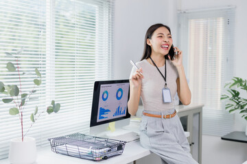 Young office worker happily talking on the phone and holding a pen while sitting on her desk, with a desktop computer displaying charts and graphs in the background, suggesting a productive workday