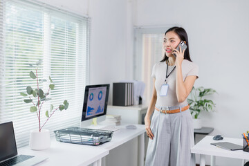 Young Asian businesswoman wearing an ID card, engaged in a phone conversation, standing in a modern office filled with computers and greenery, exuding confidence and professionalism