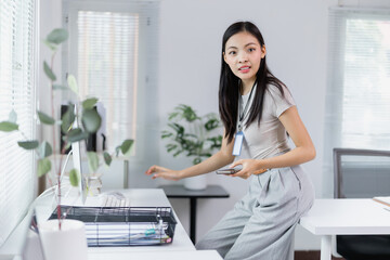 Young Asian businesswoman wearing lanyard with id card is using smartphone and working with desktop computer in modern office, business and technology concept