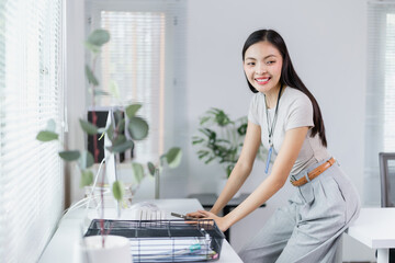 Young Asian businesswoman smiling while leaning on her desk in a modern office workspace with plants and natural light, showcasing a positive and productive work environment