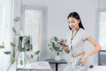 Young Asian businesswoman using mobile phone in modern office with computer and plants, she is wearing casual clothes and standing near her desk