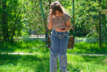 Happy girl on swing, teenage girl in headphones listening favorite  music in smartphone, vlogging, enjoying summer time. Life Events. Candid, real people