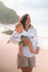 Portrait of a young woman walking on the beach with a baby in her arms. She hugs and kisses him on...