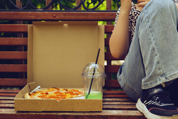 Cute girl having fun sitting on bench in the city park,  eating pepperoni pizza, drinking lemonade with ice. Happy teen in the public park, street food, having lunch. Activity and leisure travel.