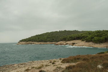 Panoramic view of a rocky Adriatic coastline covered with dense Mediterranean pine forest under a moody sky. Calm sea water with a few floating buoys.