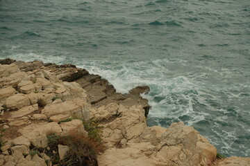 Jagged limestone rocks meet the rough Adriatic Sea as waves crash into the rugged shoreline. The dynamic contrast of textured stone.