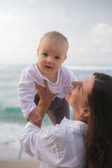 Close-up portrait of a baby with blue eyes being held by his mother against ocean. The mother is...