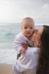 Close-up portrait of a baby with blue eyes being held by his mother against ocean. The mother is...