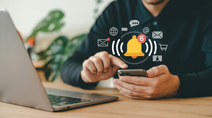 A man is using a laptop and a cell phone. The cell phone has a bell on it and the man is touching it. The laptop is open and has a green plant on it. Concept of staying connected