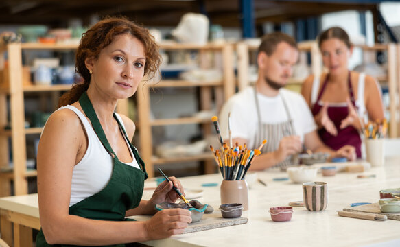 Female apprentice, is sitting near desktop and coloring clay product. In background, lovers of applied art complete design of bowl made of clay, apply pattern with brush in presence of teacher
