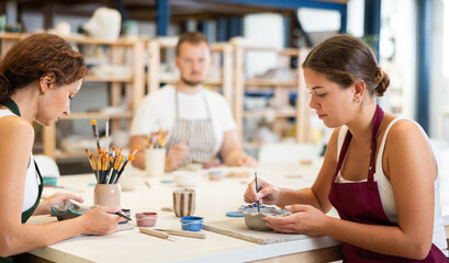 In pottery workshop, two girls and guy are sitting at table, busy coloring products made of raw...