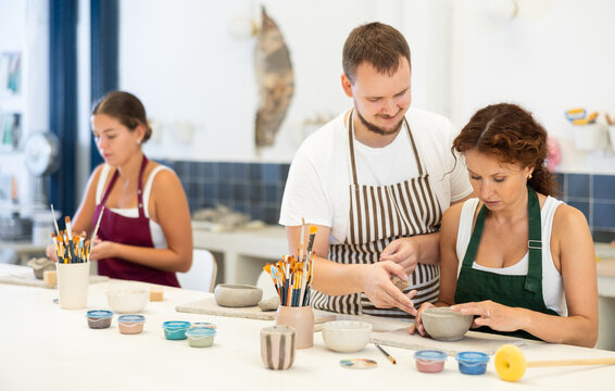 In pottery workshop, apprentice female students sitting at table, engaged in making products from raw clay in presence of teacher. Visitors to clay craft workshop work under guidance of male mentor