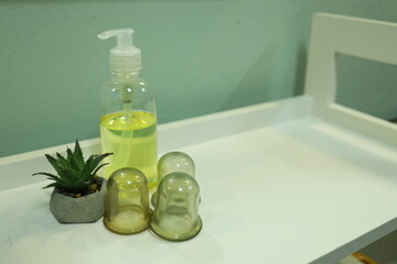 A clear bottle filled with yellow oil sits next to three green glass cups on a white table. A small...