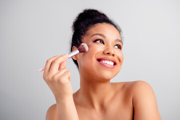 Beautiful young woman smiling and applying makeup using a brush against a grey background